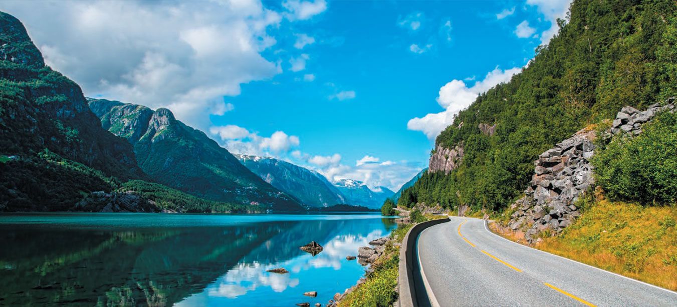 A scenic road winds along the edge of a reflective lake in Norway, with steep, tree-covered mountains rising on either side.