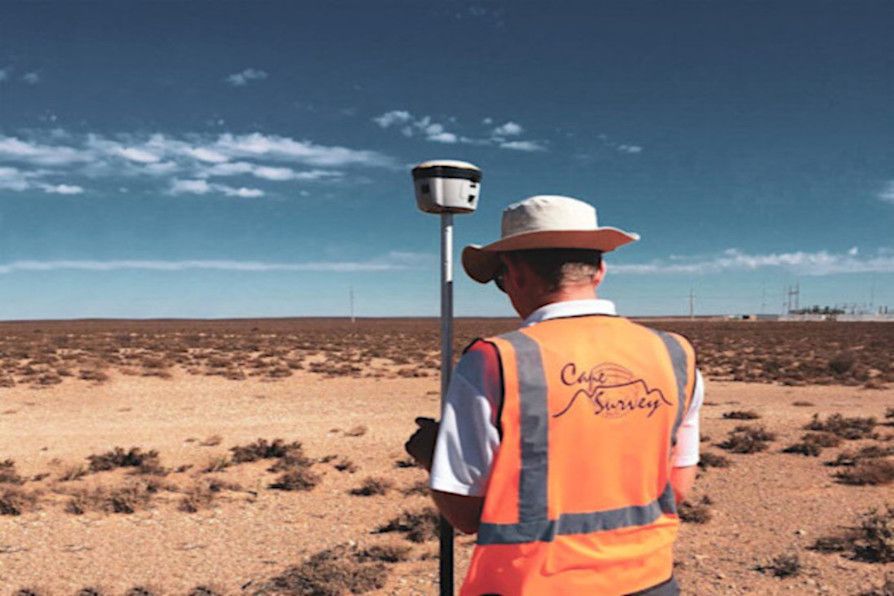Surveyor in a high-visibility vest uses a GPS device in a vast, arid landscape under a blue sky.