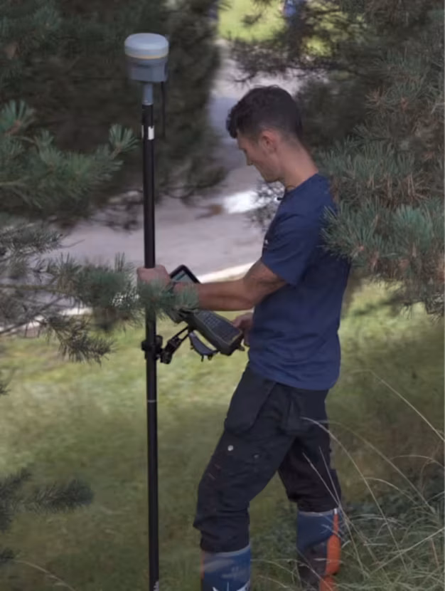 A surveyor holding a GNSS pole with a data collector attached, standing in a grassy area with trees.