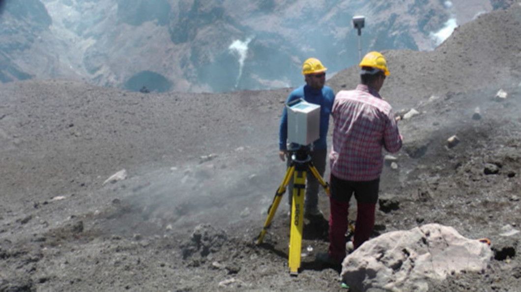 Two men in yellow hard hats stand on a rocky, desolate terrain, one operating a Trimble laser scanning device mounted on a tripod.
