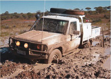A white 4x4 truck is stuck in thick mud, with its tires submerged and mud splattered all over the vehicle.