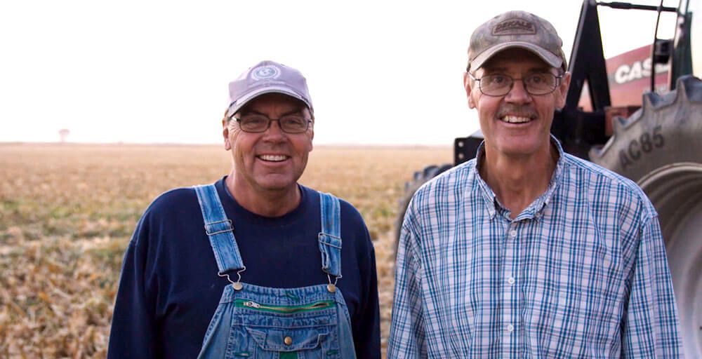 Two farmers smile at the camera in front of a tractor and a harvested field.