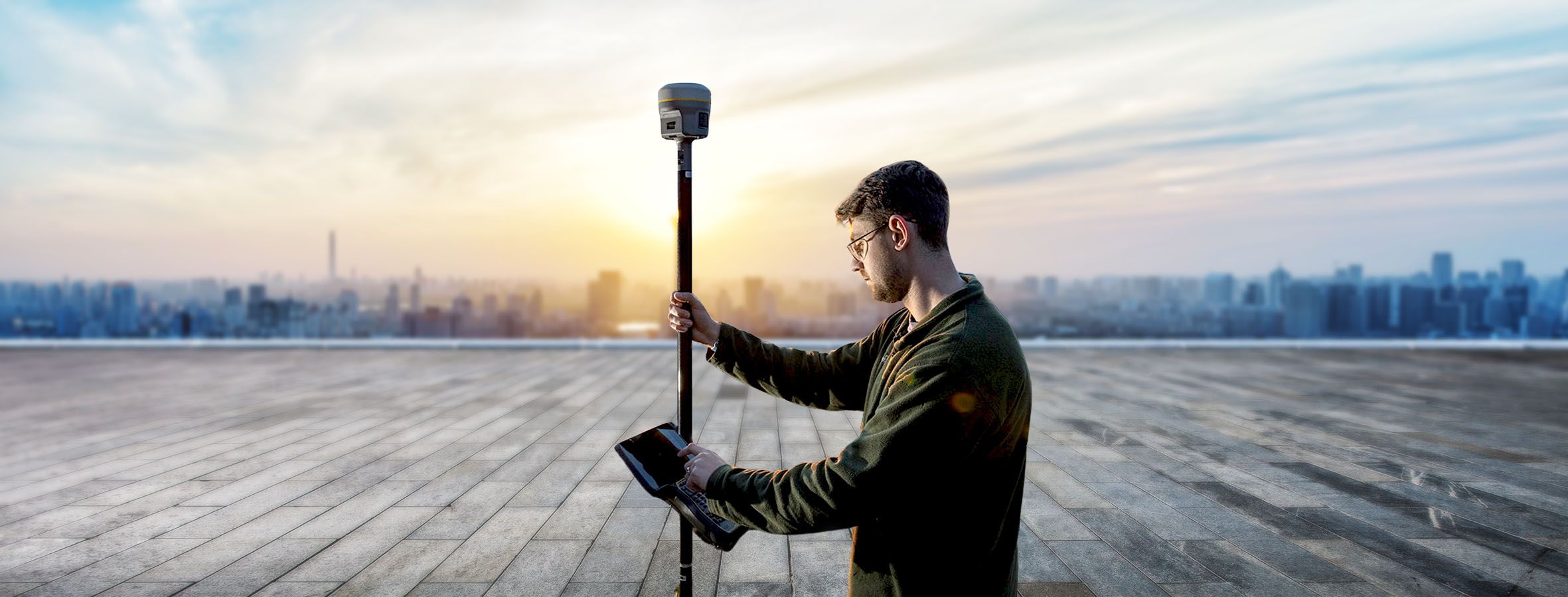 A surveyor on a rooftop holds a GPS antenna and tablet computer with a cityscape in the background at sunset.